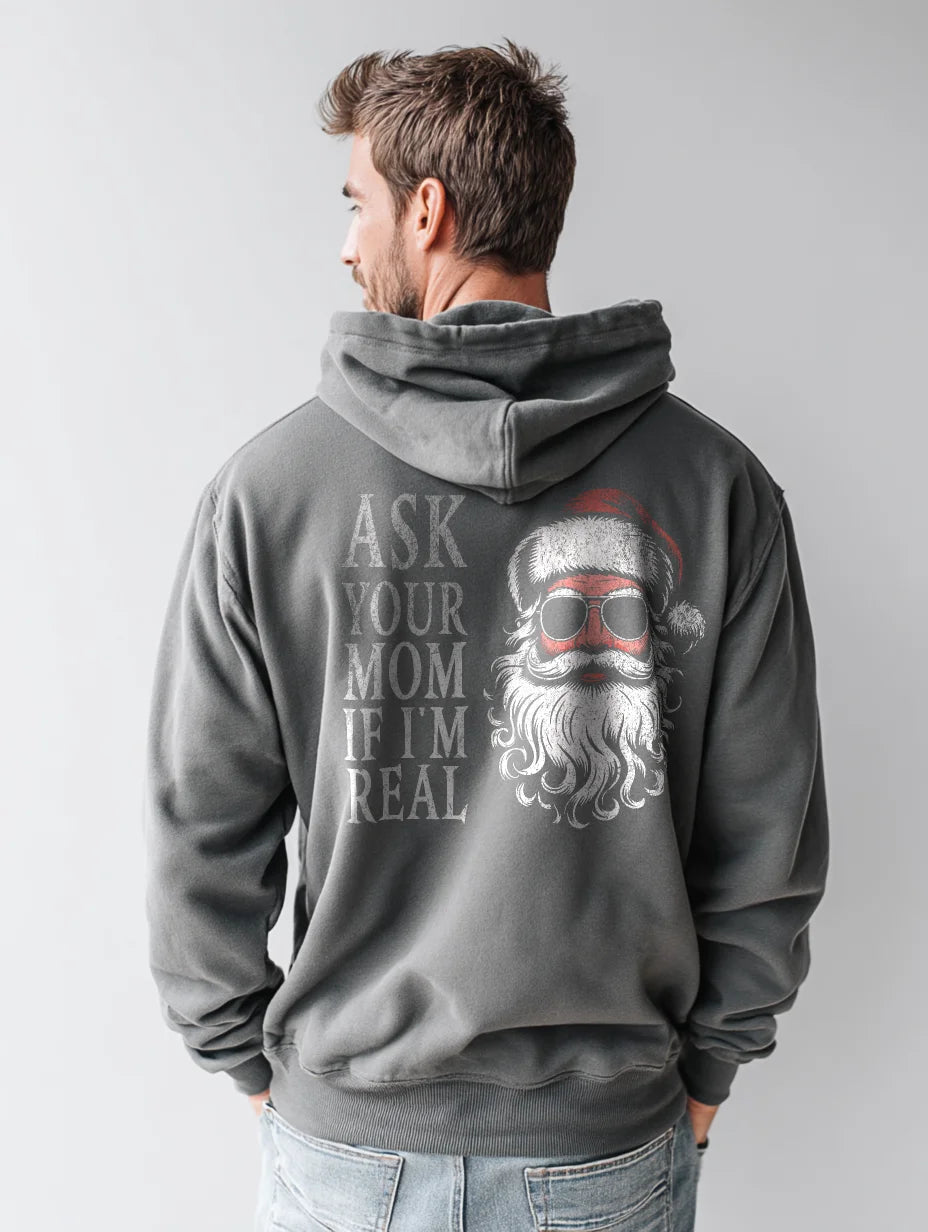 Charcoal men’s hoodie with large vintage Santa face and sunglasses graphic on the back and distressed text that reads “Ask Your Mom If I’m Real,” modeled by a man standing in a studio with a gray background.
