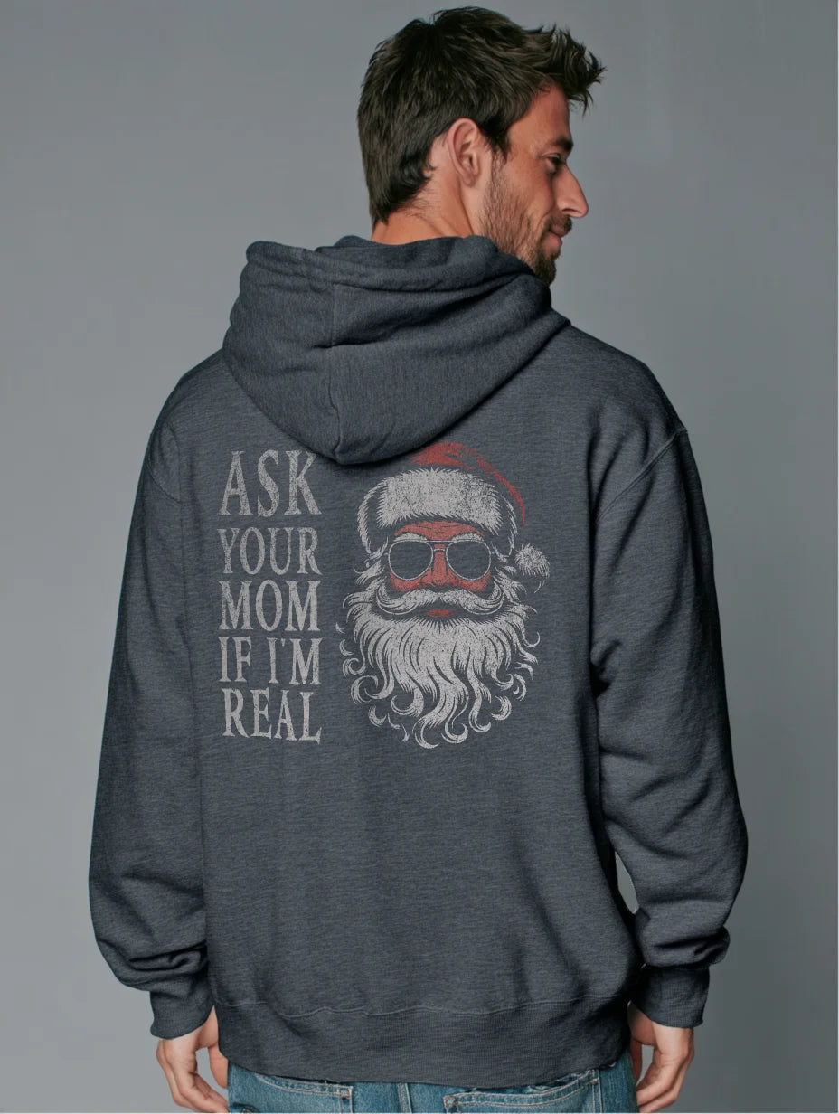 Dark Heather men’s hoodie with large vintage Santa face and sunglasses graphic on the back and distressed text that reads “Ask Your Mom If I’m Real,” modeled by a man standing in a studio with a gray background.