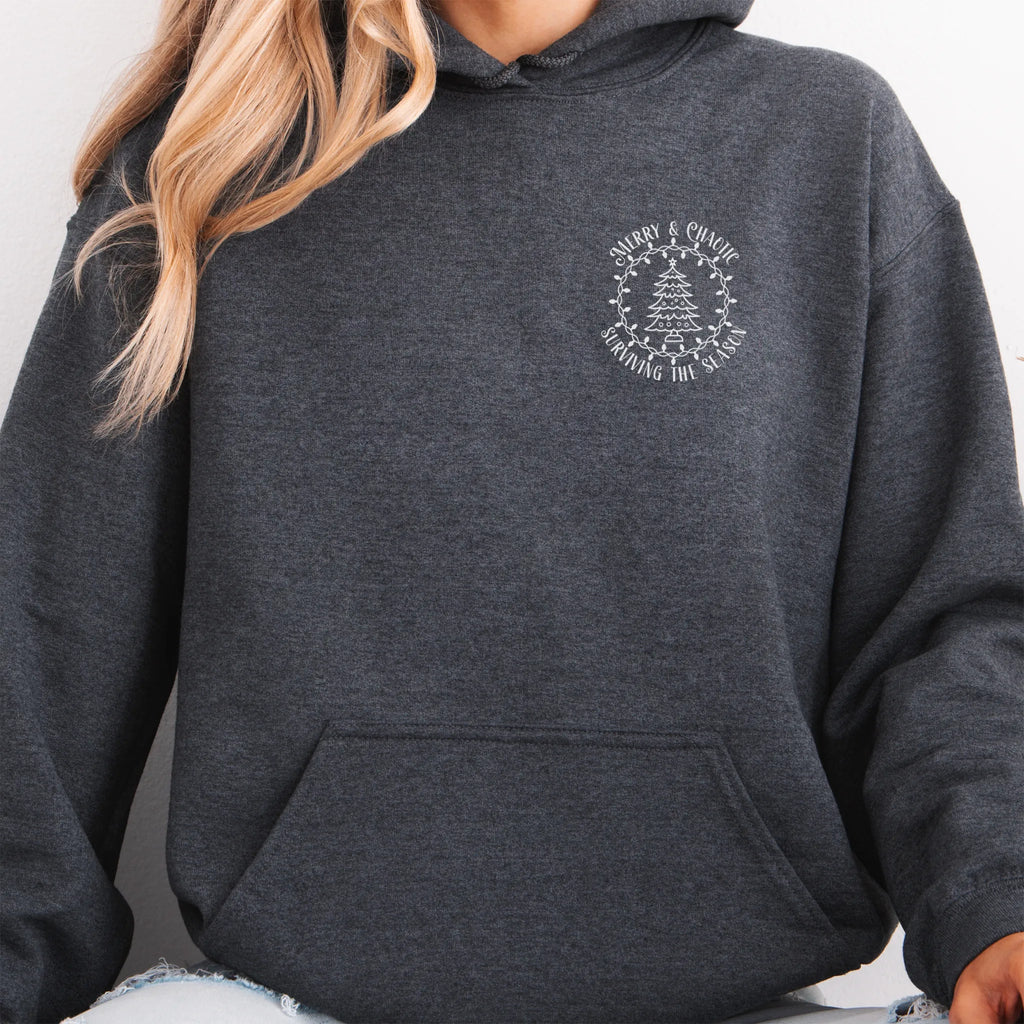 Dark Heather women’s hoodie left chest badge text “Merry & Chaotic — Surviving the Season”
