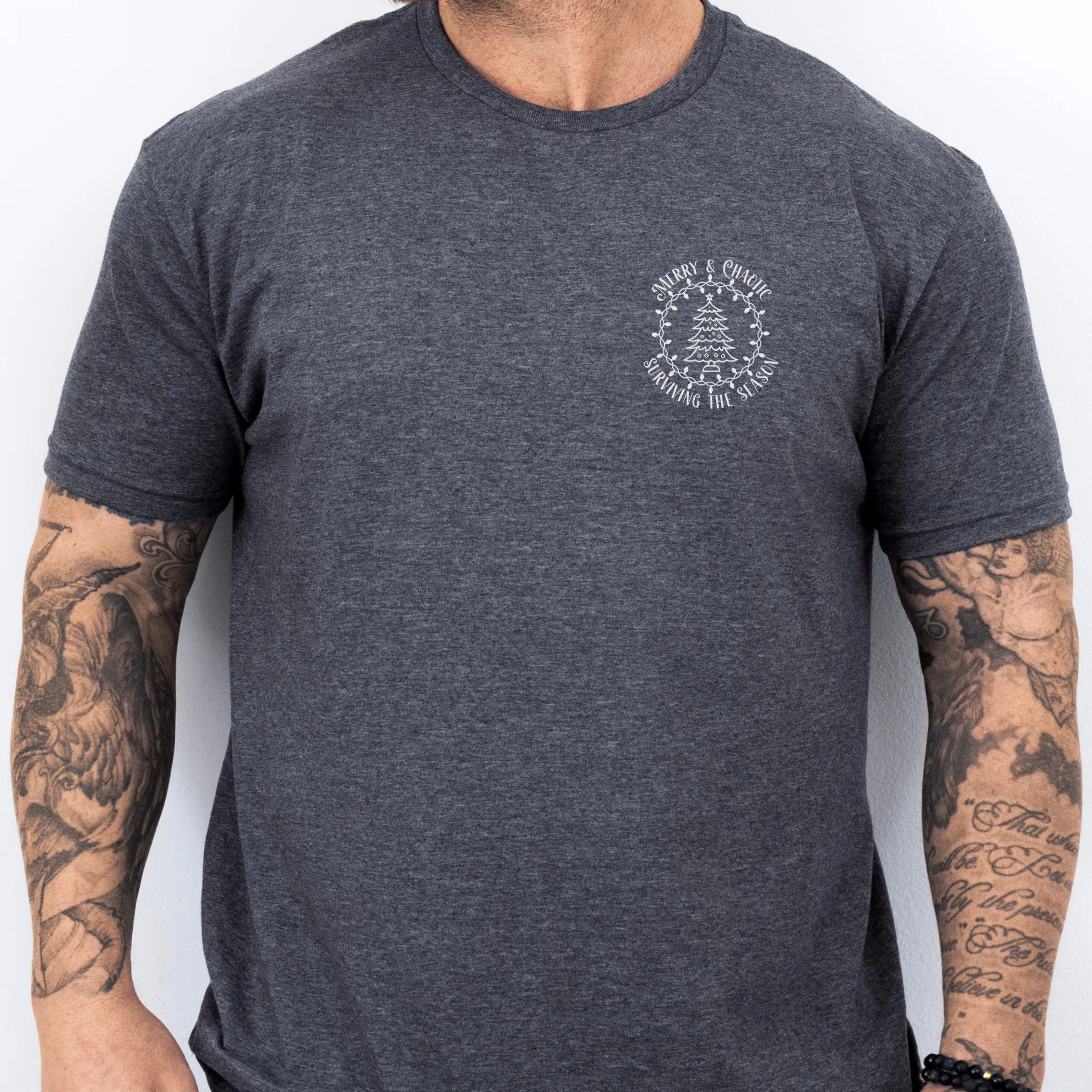 Dark Heather Oakley June tee left chest badge text “Merry & Chaotic — Surviving the Season” with Christmas tree