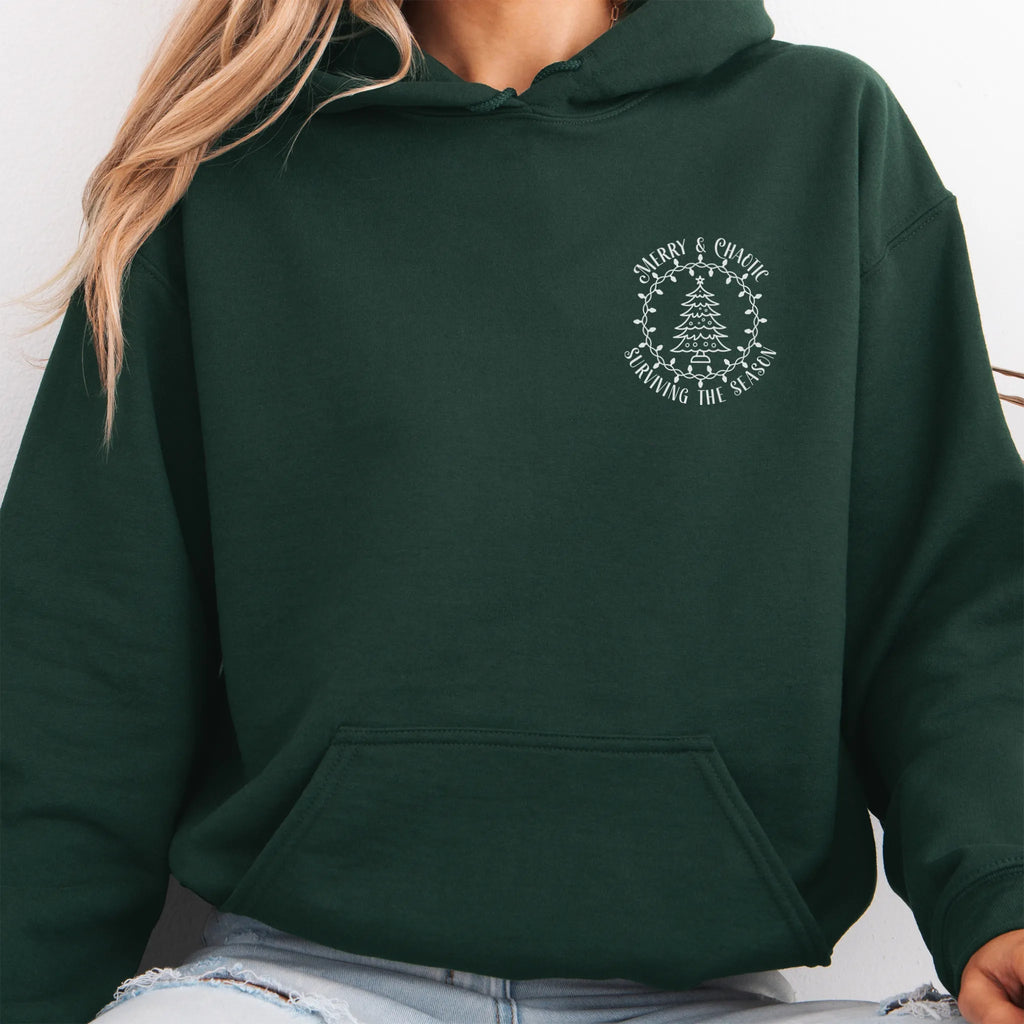 Forest Green women’s hoodie left chest badge text “Merry & Chaotic — Surviving the Season”