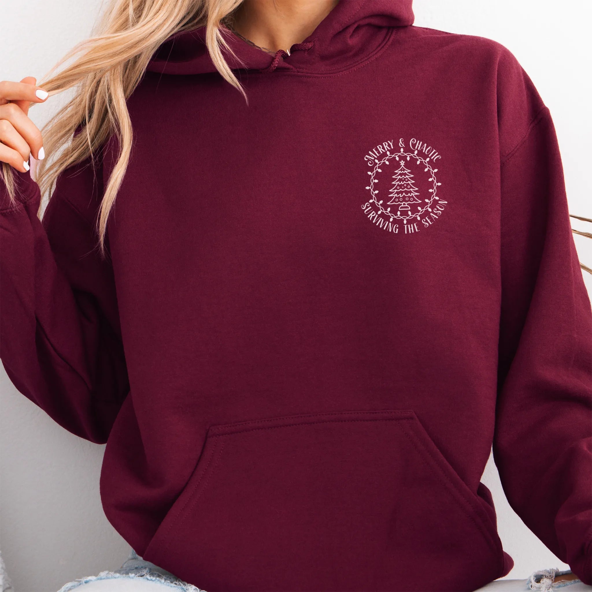 Maroon women’s hoodie left chest badge text “Merry & Chaotic — Surviving the Season”