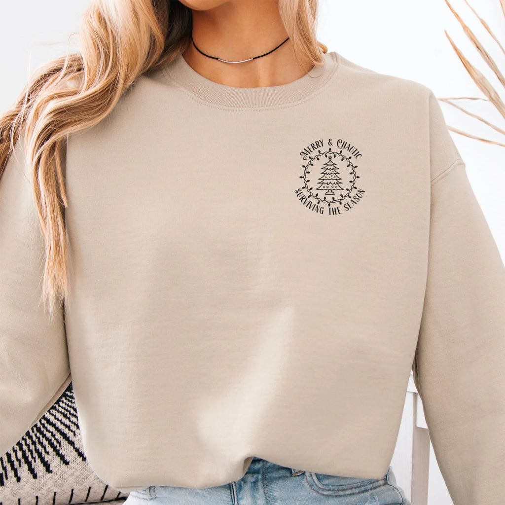 Sand women’s sweatshirt left chest badge text “Merry & Chaotic — Surviving the Season”