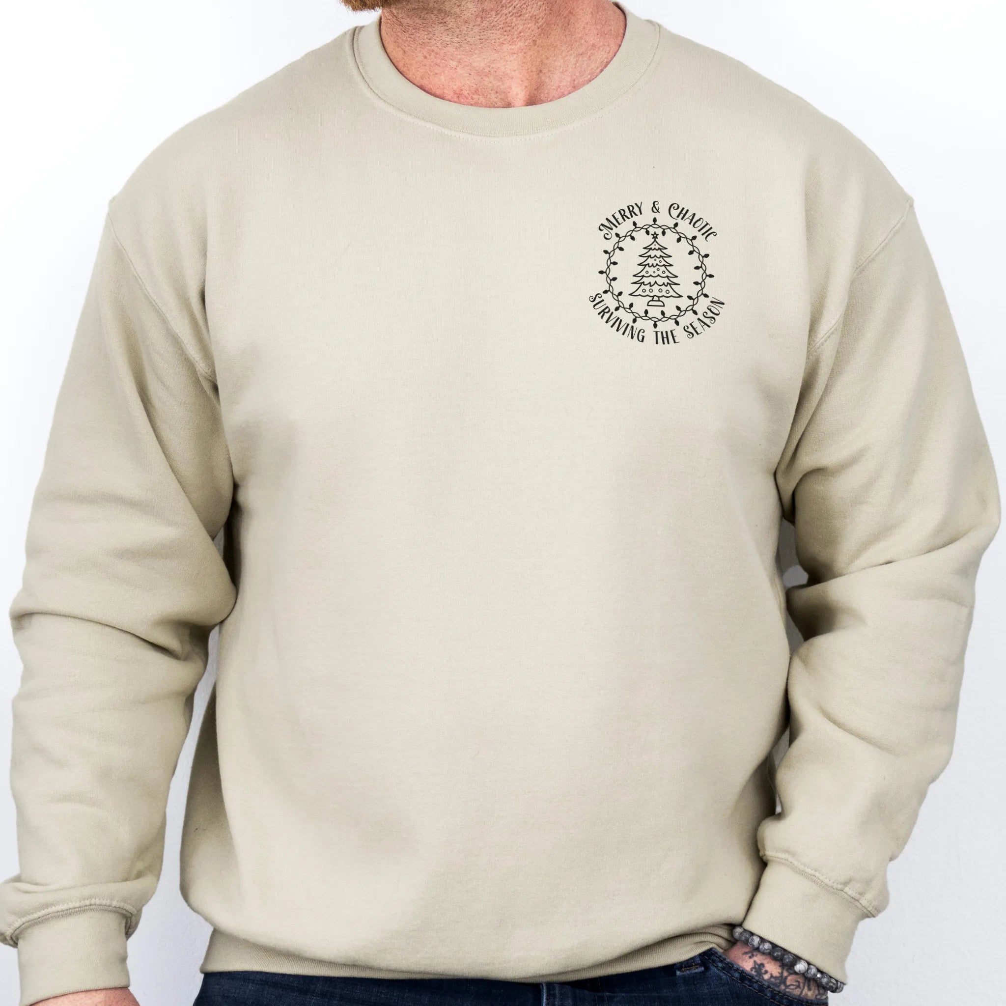 Sand men’s sweatshirt left chest badge text “Merry & Chaotic — Surviving the Season”